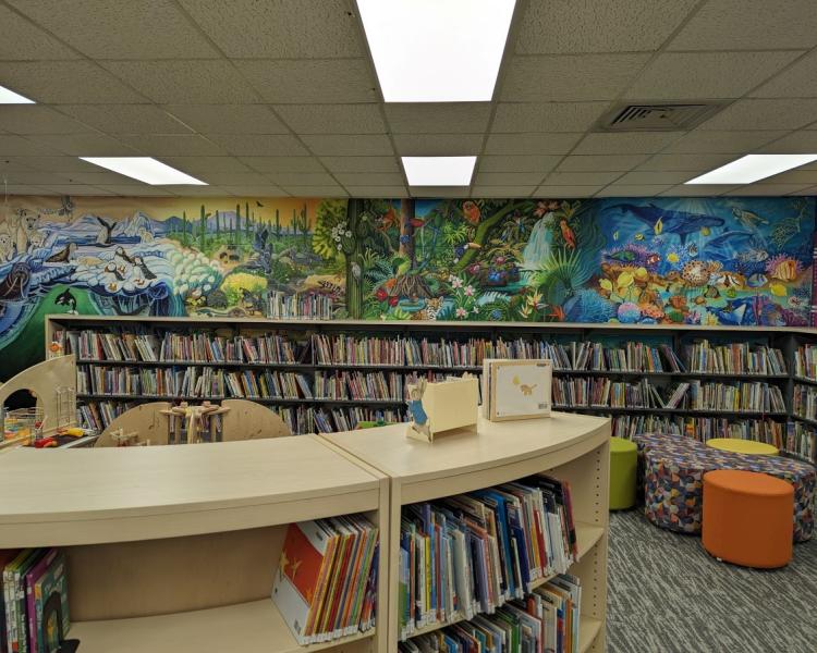 Photo of colorful murals over low shelves in the children's library; murals depict animals in ocean, forest, desert, and arctic environments